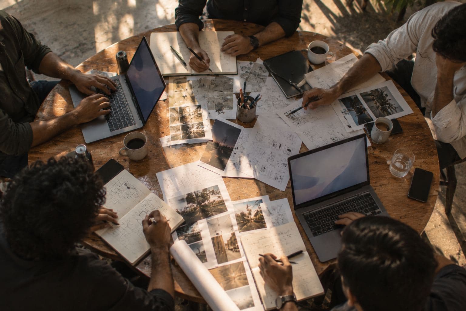 A round table from above with five people's hands, laptops and sketches.